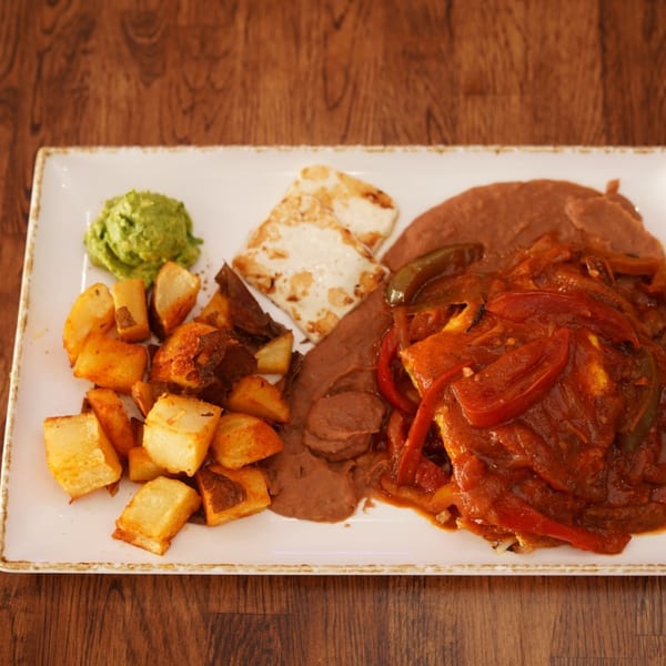 Mexican-style platter with grilled chicken, refried beans, roasted potatoes, guacamole, and sautéed peppers and tomatoes on a white plate.