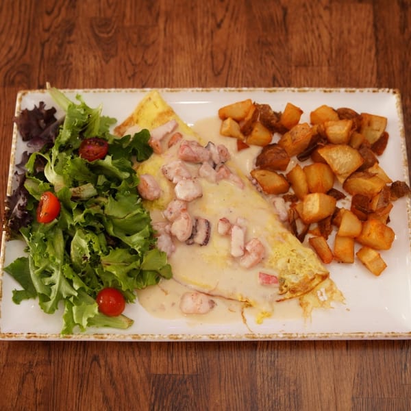 Seafood-stuffed omelet with creamy sauce, roasted potatoes, and mixed green salad on a rectangular plate.