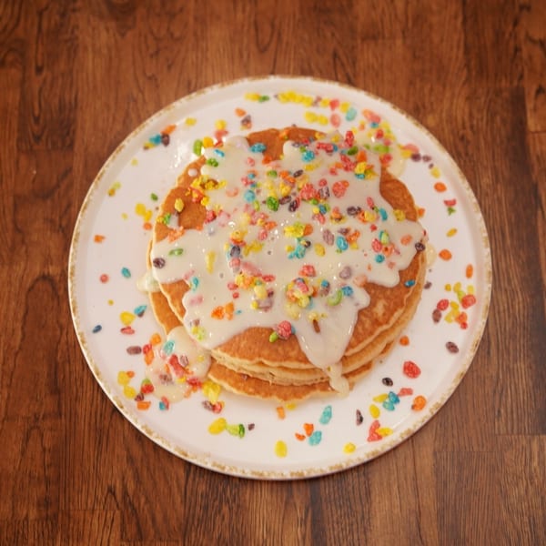 Stack of pancakes topped with white icing and colorful cereal pieces on a plate atop a wooden table.