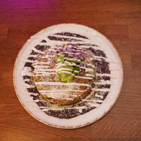 Golden enchilada topped with guacamole, red onion, cilantro, and cream sauce, served over black beans on a ceramic plate.