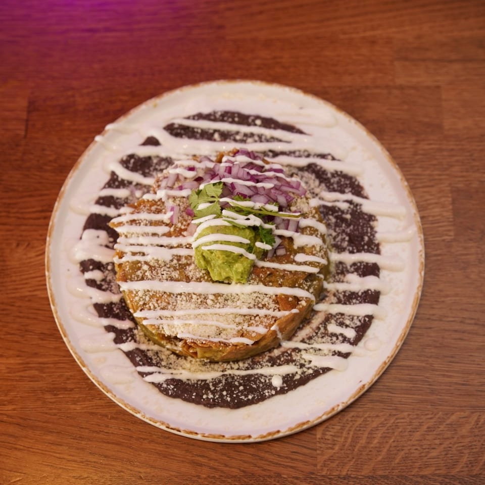 Golden enchilada topped with guacamole, red onion, cilantro, and cream sauce, served over black beans on a ceramic plate.