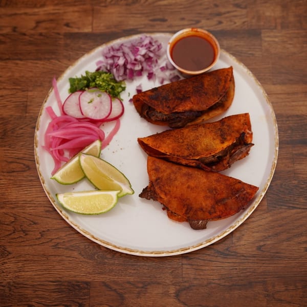 Three crispy birria tacos with lime wedges, pickled onions, radishes, cilantro, and consommé on a plate.
