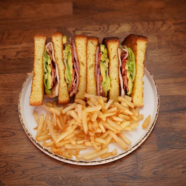 Plate of toasted club sandwiches with lettuce, tomato, avocado, and bacon alongside golden French fries on a wooden table.