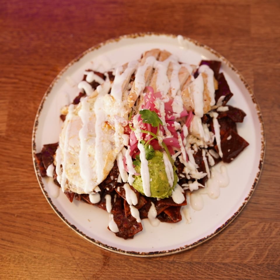 Loaded plate of saucy nachos topped with sliced chicken, guacamole, pickled onions, cilantro, sesame seeds, and crema.