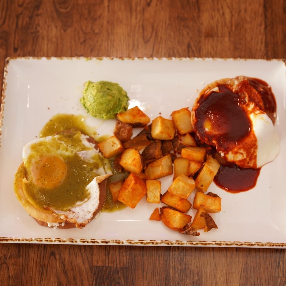 Plate of eggs Benedict with green and red sauces, guacamole, and roasted diced potatoes on a wooden table.