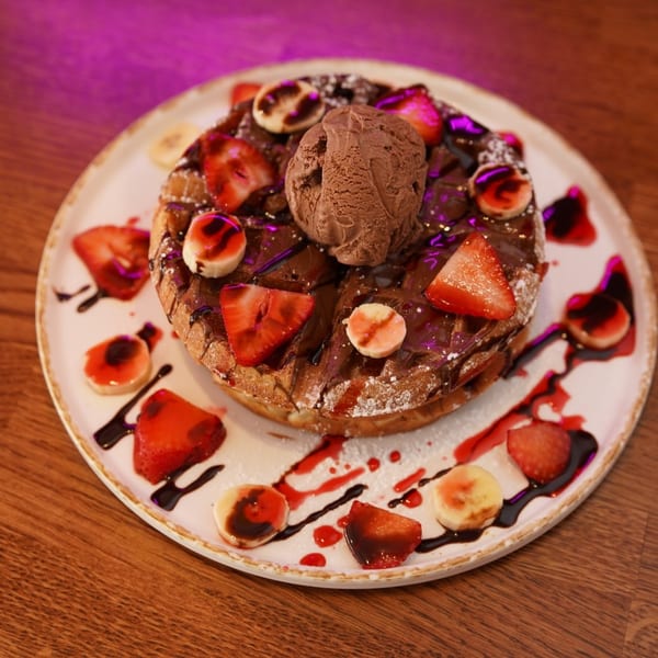 Chocolate waffle topped with ice cream, strawberries, bananas, powdered sugar, and chocolate and berry sauces on a plate.