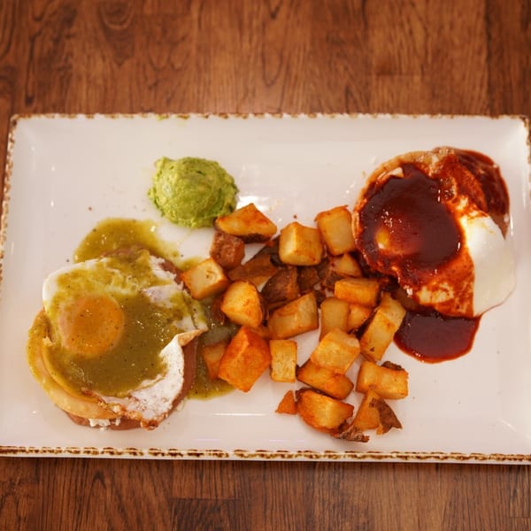 Plated breakfast featuring eggs with green salsa, roasted potatoes, avocado, and red-sauced enchiladas on a rectangular white dish.