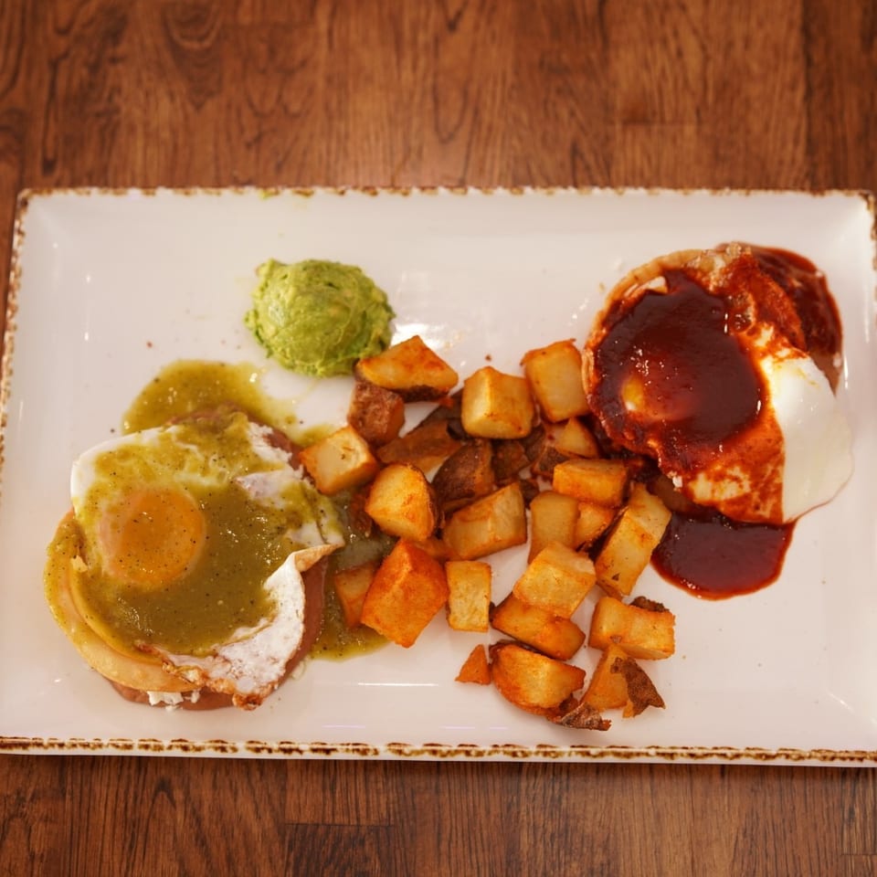 Plated breakfast featuring eggs with green salsa, roasted potatoes, avocado, and red-sauced enchiladas on a rectangular white dish.