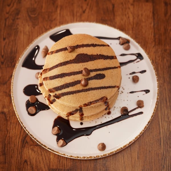 Stack of pancakes drizzled with chocolate sauce, topped with nuts, and served on a white plate over a wooden table.