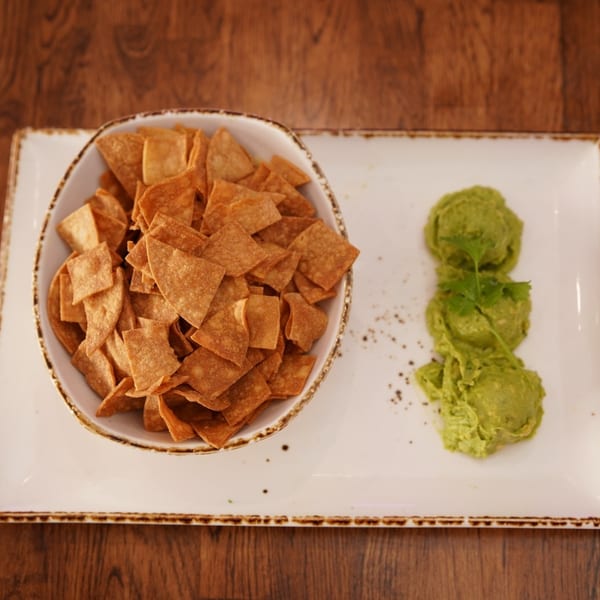 Bowl of crispy tortilla chips with three scoops of guacamole on a rectangular white plate atop a wooden table.
