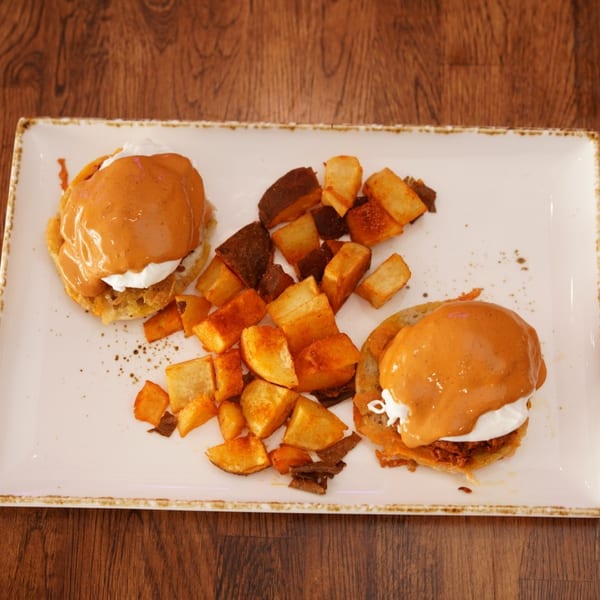 Two orange-sauce-topped sandwiches with creamy filling and roasted potato cubes on a rectangular plate atop a wooden table.