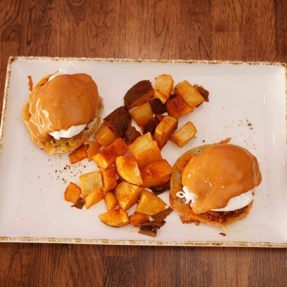 Two orange-sauce-topped sandwiches with creamy filling and roasted potato cubes on a rectangular plate atop a wooden table.
